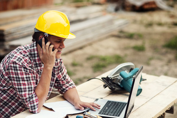 Photo of a construction worker with hard hat reviewing laptop and other materials at job site